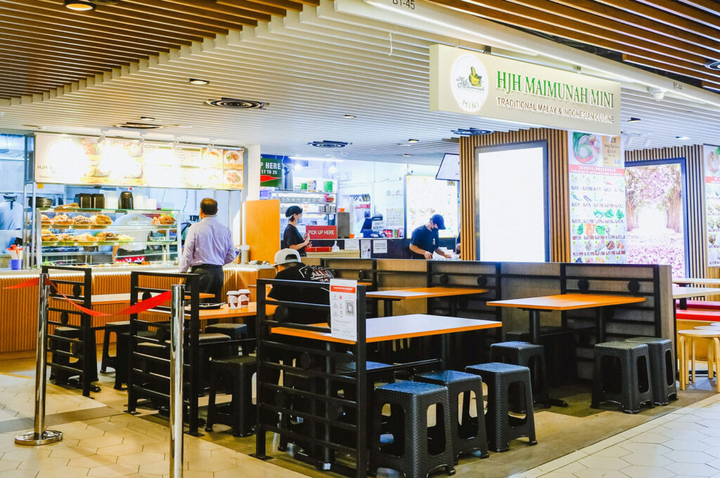 Interior view of Hjh Maimunah Mini food stall in a Singapore food court, specializing in Traditional Malay and Indonesian Cuisine. Diners are seated at black and orange tables and stools.