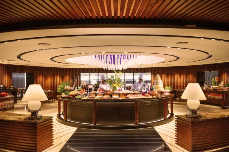 Upscale buffet restaurant with circular food display, chefs preparing gourmet dishes, modern wooden interiors, and elegant lighting design.