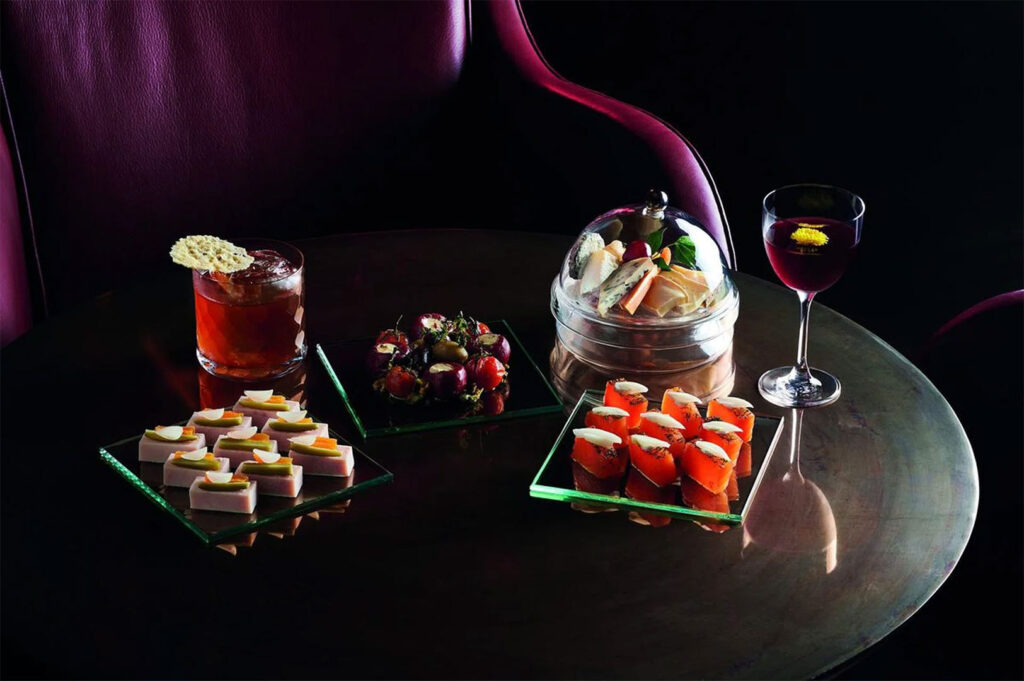A selection of sophisticated bar snacks, including small savory bites and a dome-covered plate, paired with two cocktails on a dark, reflective table in an upscale bar.