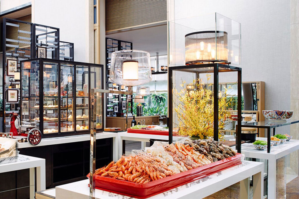An elegant indoor buffet station in a bright setting, displaying fresh chilled seafood like prawns and oysters on a red tray, with a separate glass display for desserts in the background.