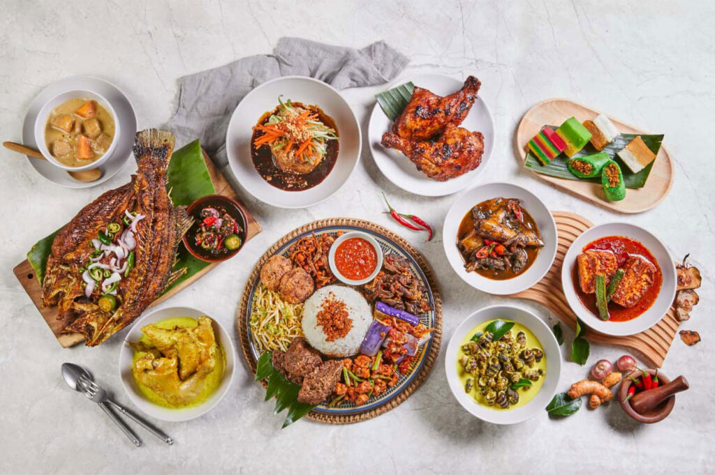 Overhead shot of a large, diverse spread of Malay and Indonesian Nasi Padang dishes on a white marble table, including a platter of rice and various meat/vegetable sides, whole fried fish, chicken, bowls of curries, and colorful kuih (desserts).