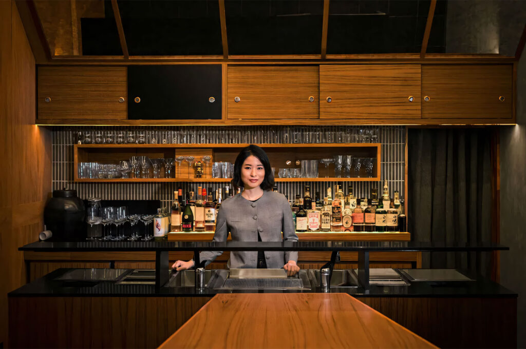 A female bartender stands professionally behind a sleek, modern Japanese-style bar counter with wooden accents and shelves displaying various vintage spirits and glassware.