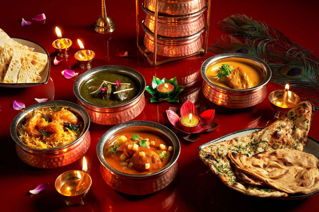 A festive table setting featuring various Indian curries served in copper bowls (karahi) with naan and papadums. The setting is illuminated by small tea lights and decorated with rose petals and a peacock feather, suggesting a Diwali celebration.