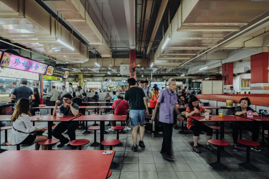 Busy indoor Singapore hawker center food hall with red tables and stools. People are queuing at stalls and sitting down to eat, showing a typical casual dining environment.
