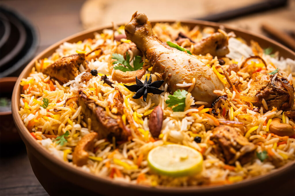 A close-up shot of a large bowl of fragrant Chicken Biryani. The dish consists of long-grain rice, chicken pieces including a drumstick, spices, and is garnished with fresh cilantro and a lime wedge.