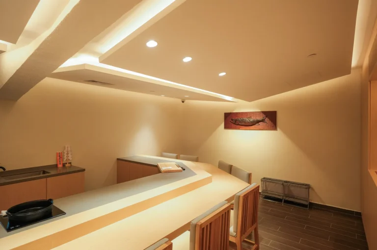 An upscale, minimalist Japanese omakase sushi bar featuring a light wood L-shaped counter, elegant recessed ceiling lighting, and clean warm interior design.