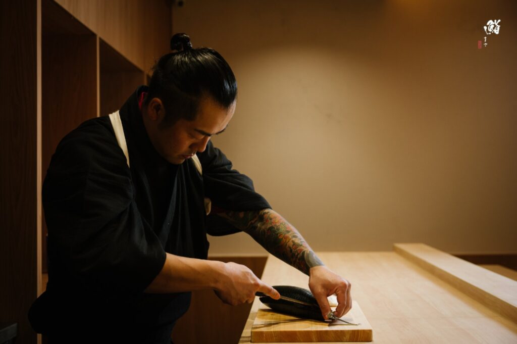 At Sushi Masa, a man with tattoos is cutting wood, demonstrating his woodworking skills and attention to detail.