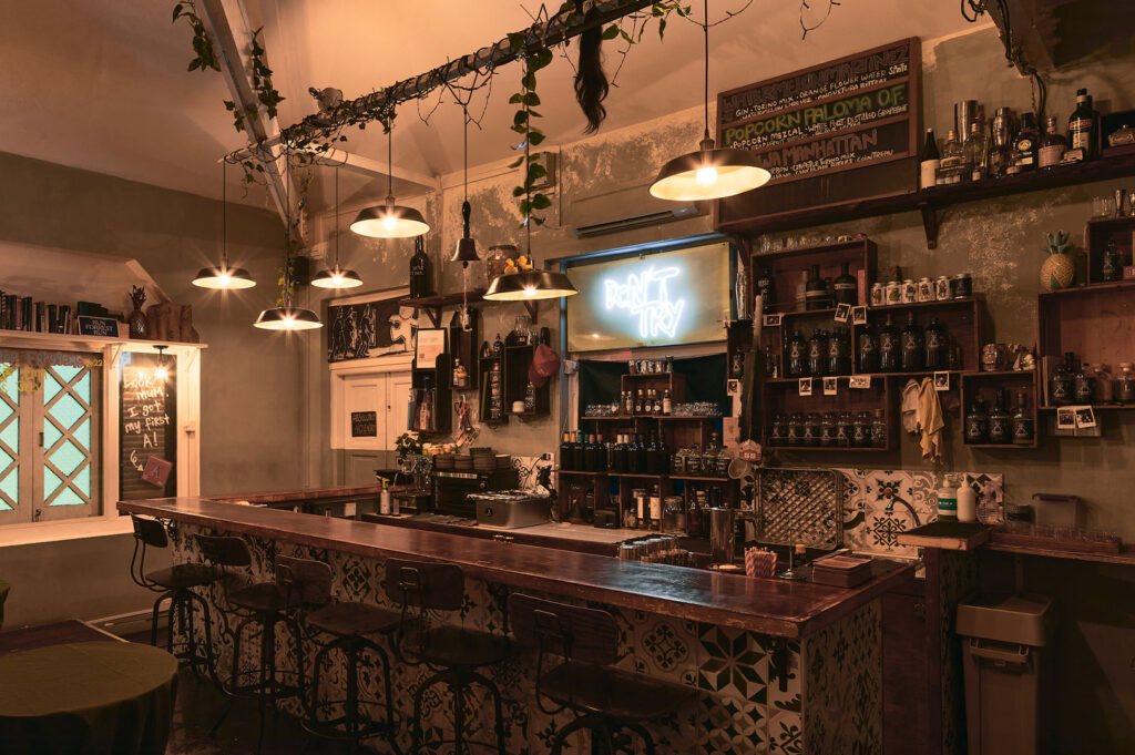 The rustic and moody interior of a cozy pocket bar, featuring a wooden counter, industrial pendant lights, and a neon sign in the background, with vines hanging from the ceiling.