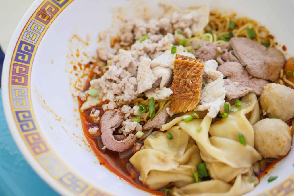 Close-up of a bowl of dry Thai-style noodles with minced pork, sliced liver, pieces of roasted pork belly, meatballs, and wontons, coated in a chili oil sauce and garnished with green onions.
