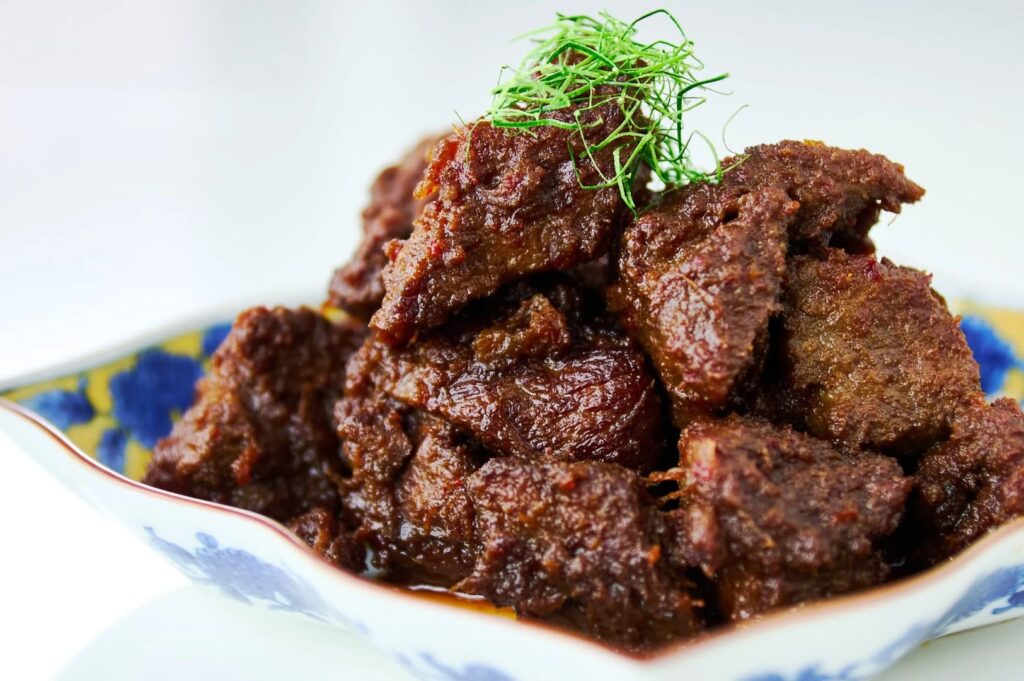A bowl of beef rendang topped with a fresh sprig of herbs, showcasing a rich and flavorful dish.