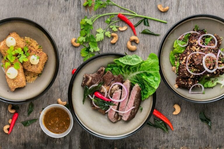 Three plates of colorful Singaporean dishes arranged on a rustic wooden table.