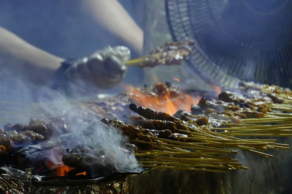 A cook prepares skewered meat with a fan in a bustling restaurant in Singapore.