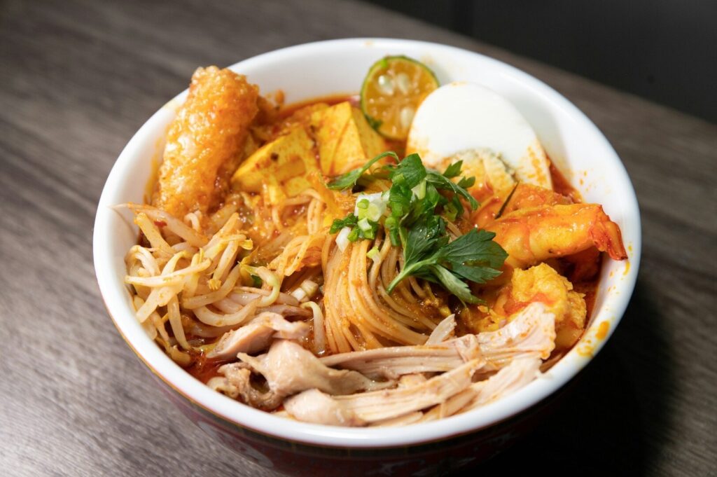 A bowl of Laksa featuring noodles, meat, and colorful vegetables in a rich broth.