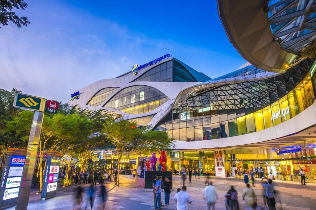People walk in front of Plaza Singapura, a modern building in a bustling city setting.