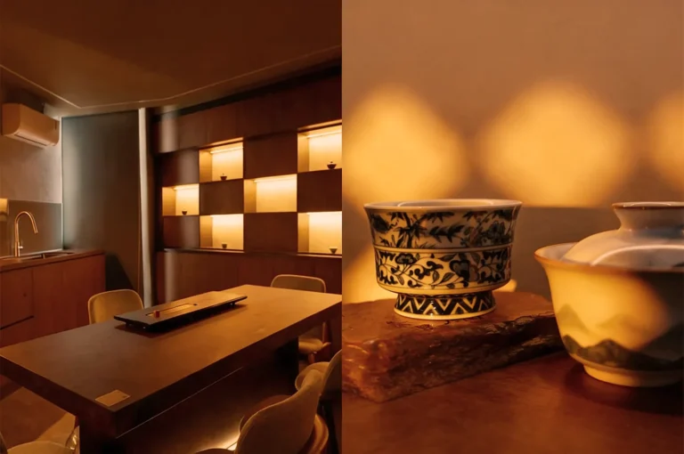 Cozy room with warm lighting features a wooden table, cushioned chairs, and an elegant shelving unit. Ceramic bowls with intricate patterns add a touch of art.