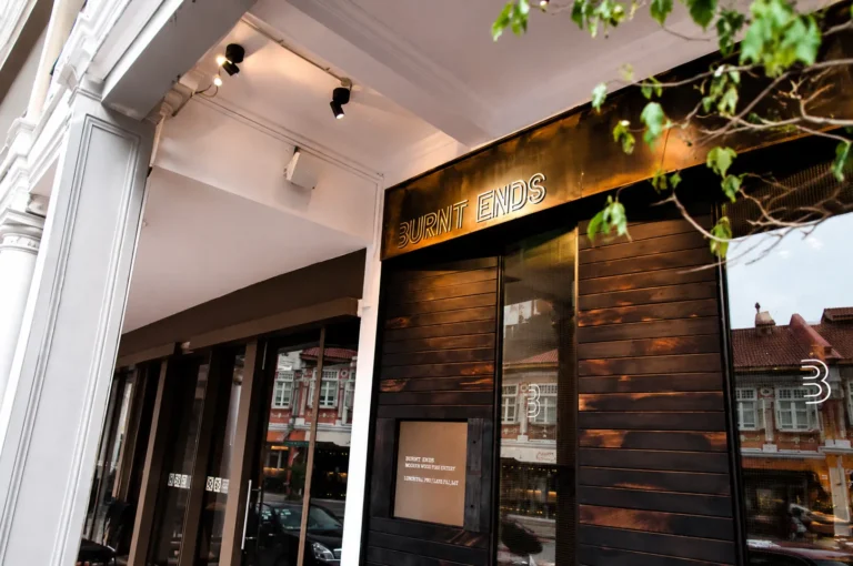 Exterior of Burnt Ends restaurant featuring a dark wooden facade and large glass windows reflecting nearby shophouses