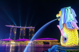 Merlion fountain illuminated at night with the Marina Bay skyline of Singapore in the background.