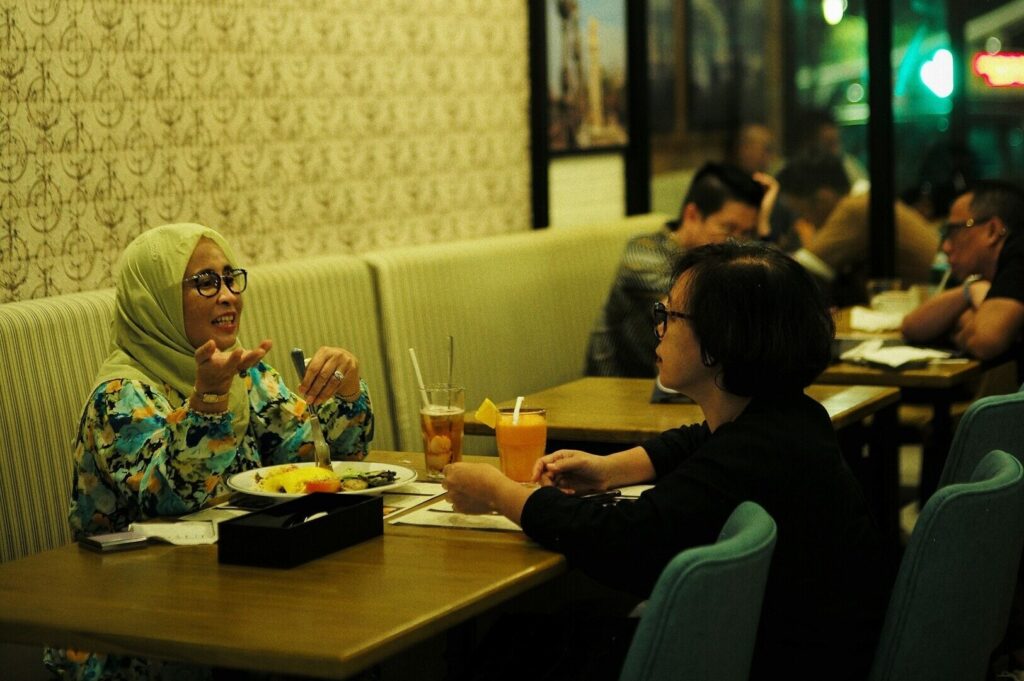 A woman wearing a hijab sits at a table with friends, sharing a meal in a lively restaurant in Singapore.