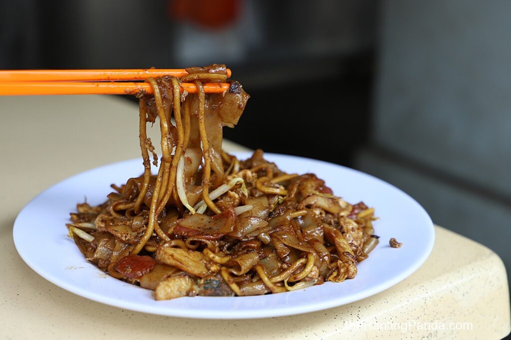 A plate of stir-fried noodles with vegetables, meat, and dark sauce, lifted by orange chopsticks, creating a savory and appetizing appearance.