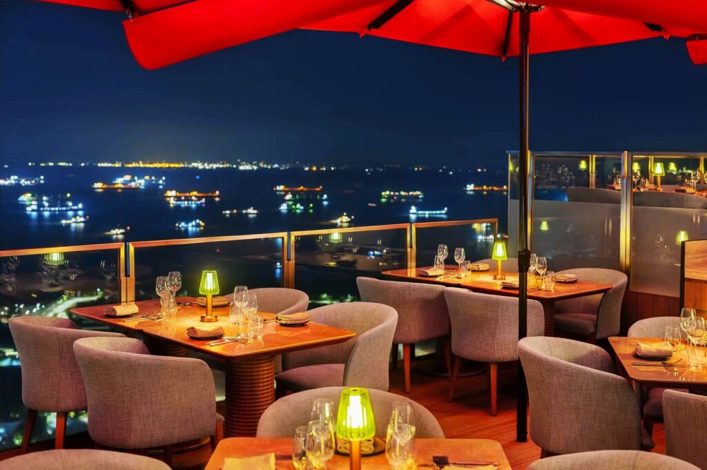Rooftop restaurant in Singapore with elegant wooden tables and gray chairs under red umbrellas at night, overlooking a cityscape of twinkling lights and a calm sea.