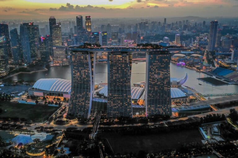 Aerial view of Singapore at dusk, featuring illuminated Marina Bay Sands with its three towers and sky park, surrounded by city skyscrapers, under a colorful sky.