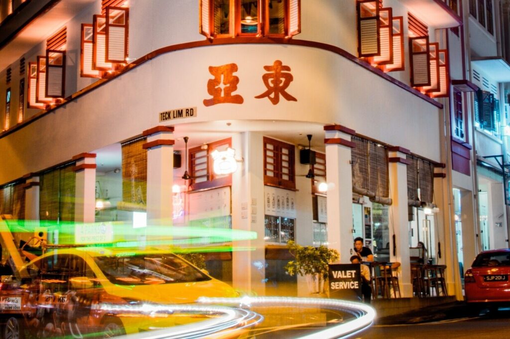 A yellow taxi drives down a street at night, with a Singaporean restaurant illuminated in the background.