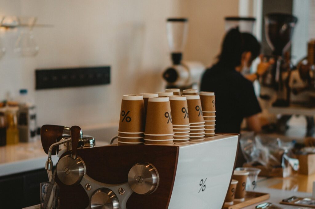 Stacked brown paper coffee cups with a percent symbol are on an espresso machine. A barista works in the blurred background, creating a cozy cafe atmosphere.