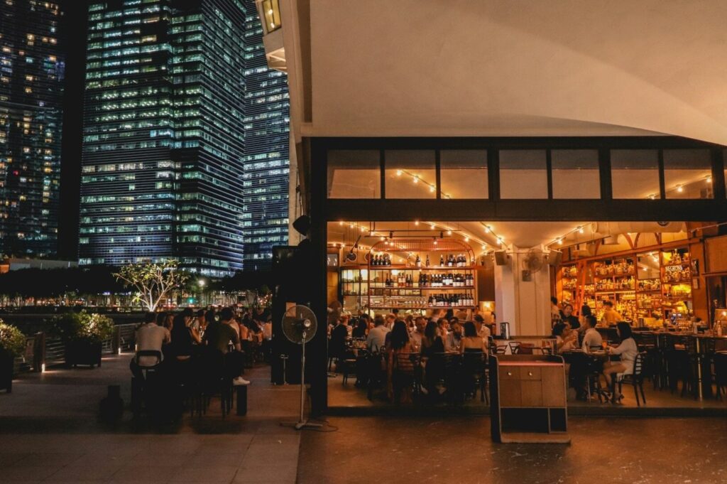 A bustling restaurant in Singapore with diners at tables, set against a backdrop of tall urban buildings.