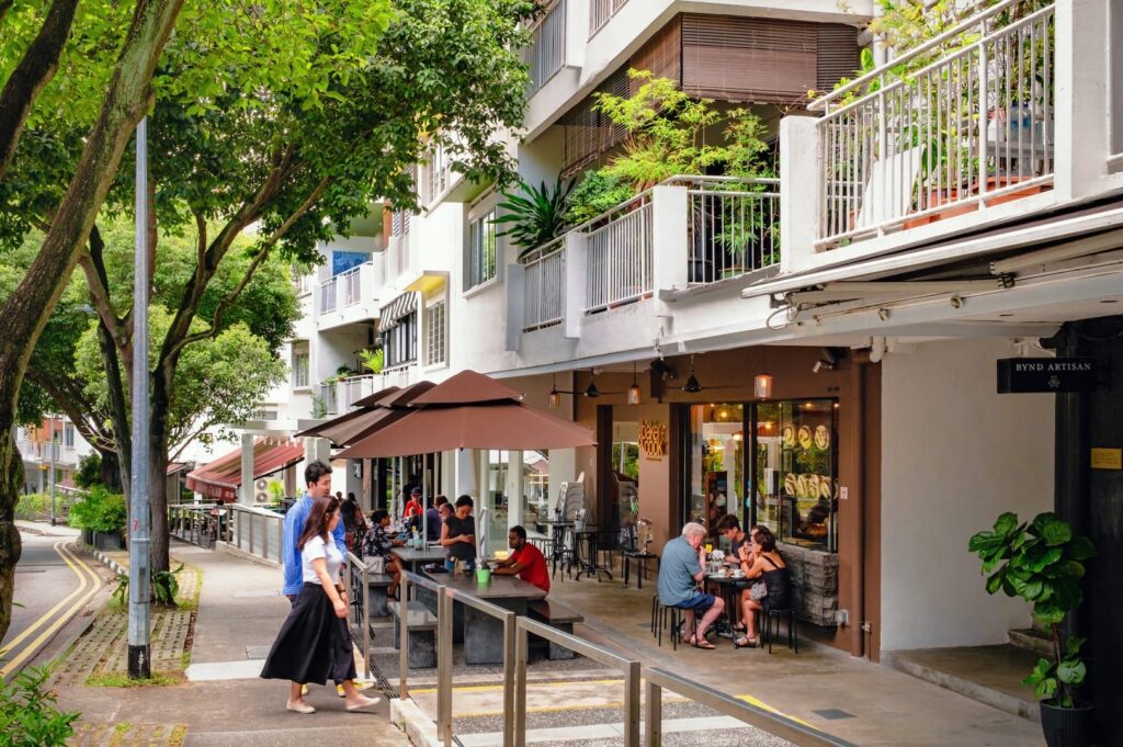 A bustling street in Holland Village, Singapore, with people dining outside a restaurant under sunny skies.