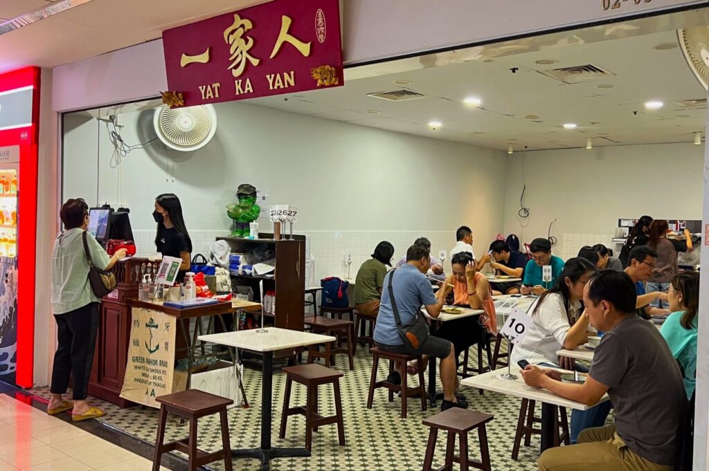A busy cafe named "Yat Ka Yan" with a red sign. Patrons seated on stools at tables engaged in conversation, creating a lively and bustling atmosphere.
