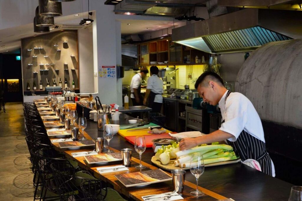 Chef in a striped apron prepares food at a modern restaurant counter. The setting features arranged dining setups and open kitchen ambiance.