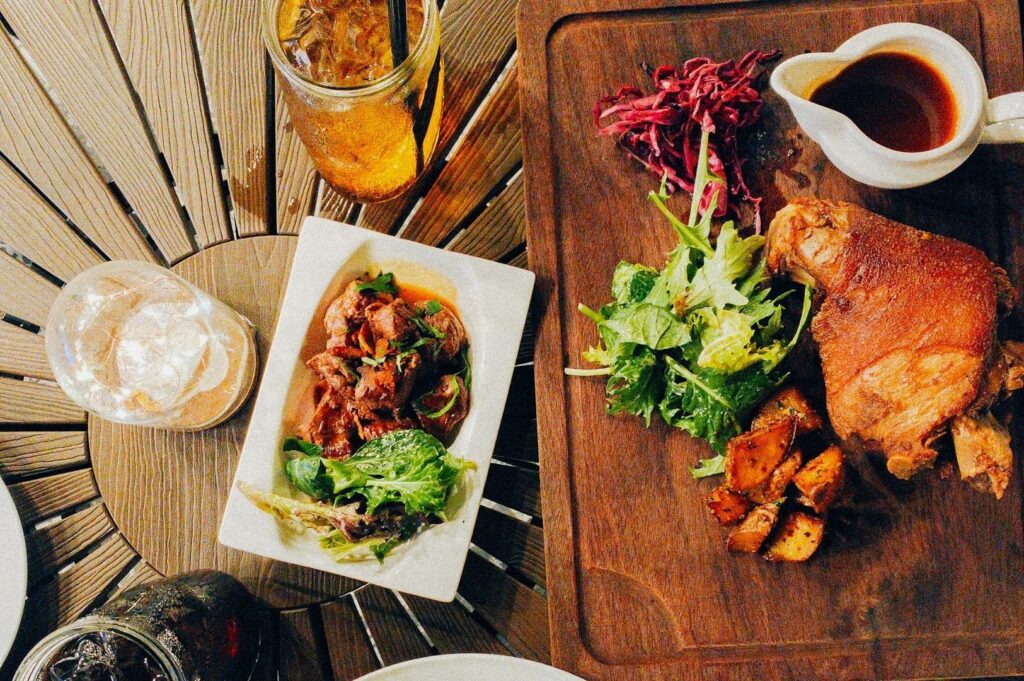 A wooden table set with iced tea, water, and soda alongside dishes of roasted meat, salad, potatoes, and red cabbage slaw. Warm, rustic ambiance.