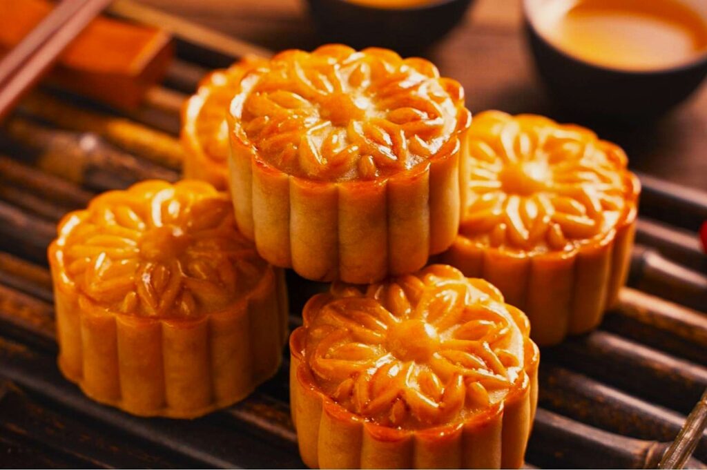Golden mooncakes with intricate floral patterns are artfully arranged on a bamboo mat. In the background, cups of tea add to the warm, inviting setting.