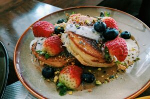 Plate of fluffy pancakes topped with fresh strawberries, blueberries, and drizzles of cream, garnished with crushed pistachios on a rustic table.