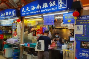 Stall with blue sign reading "Tian Tian Hainanese Chicken Rice." A person buys food from a masked vendor. Colorful bags and menu signs adorn the stall.