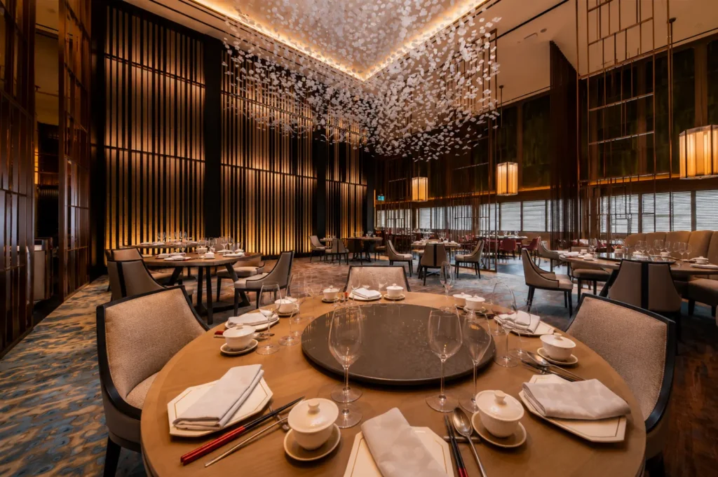 Elegant fine dining restaurant interior at Hilton Singapore with round tables, lazy susan, white porcelain settings, and dramatic floral ceiling installation