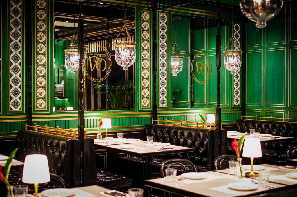 Elegant restaurant interior with deep green walls, ornate patterns, crystal chandeliers, and black leather seating. Tables are set with white lamps, conveying a classy ambiance.