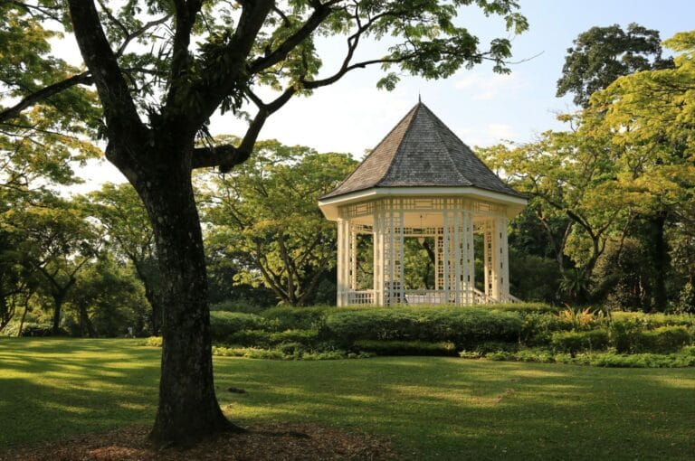 A white gazebo sits in a lush, green park surrounded by vibrant trees under a clear blue sky. The scene conveys tranquility and natural beauty.