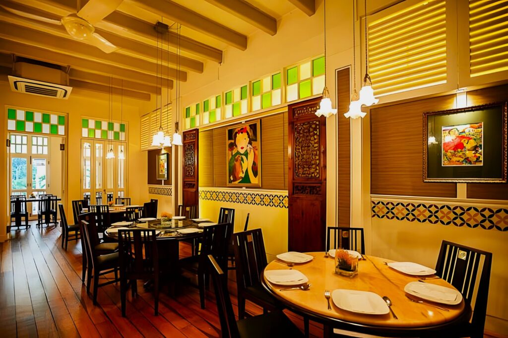 Warmly lit dining room with wooden floors, round tables, and black chairs. Colorful art and green accents adorn the walls, creating a cozy, inviting atmosphere.