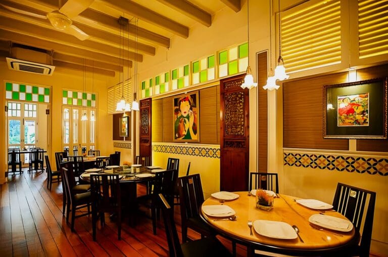 Warmly lit dining room with wooden floors, round tables, and black chairs. Colorful art and green accents adorn the walls, creating a cozy, inviting atmosphere.