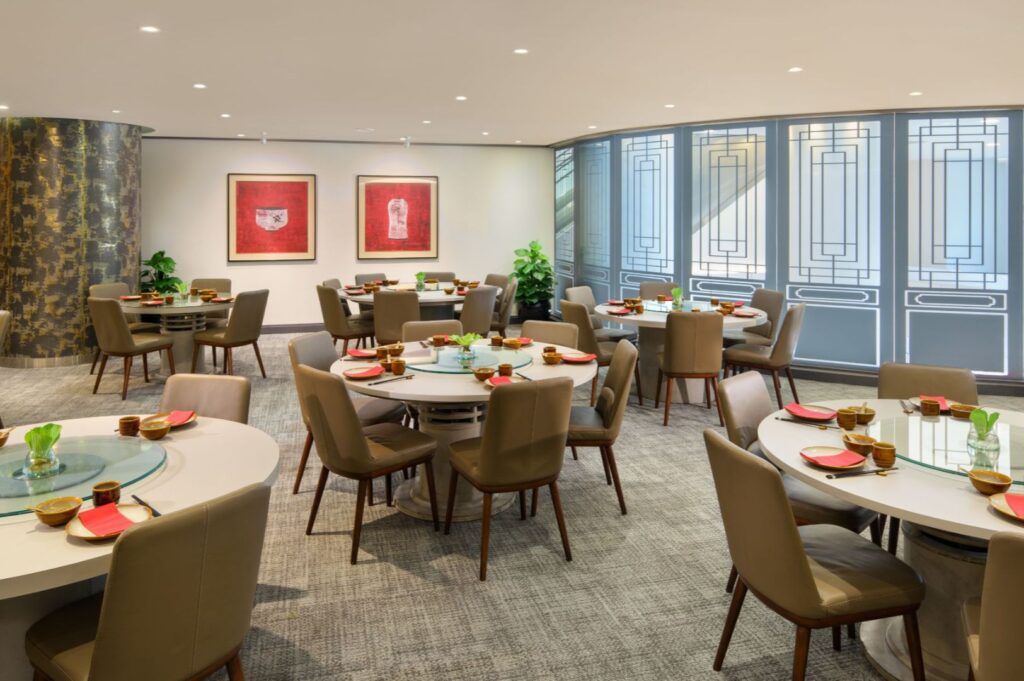 Elegant restaurant interior with round tables set for dining, surrounded by brown chairs. Red artwork and decorative panels adorn the walls, creating a sophisticated ambiance.