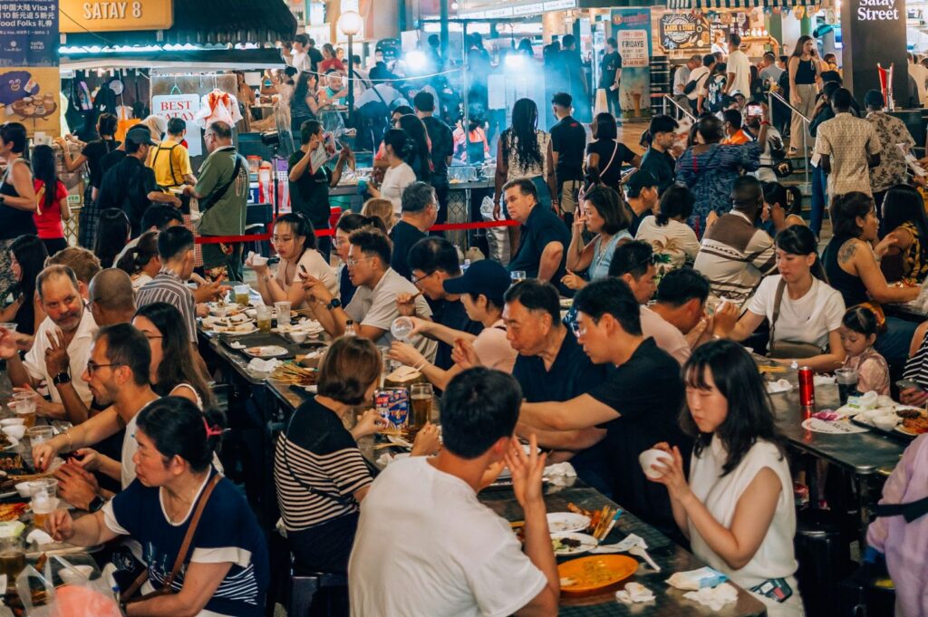 A bustling night market scene with people crowded around tables enjoying diverse street food. Vibrant atmosphere with warm lighting and lively conversations.
