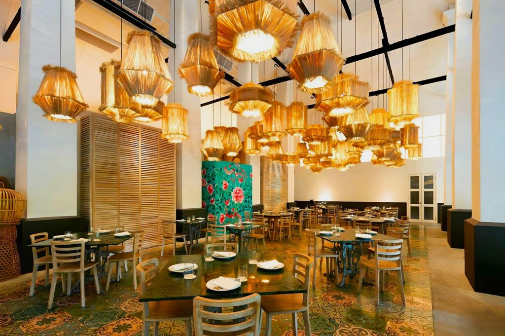 Bright, spacious dining area with colorful tiled floor and numerous warm, wooden light fixtures hanging from the ceiling. Tables with neatly set plates, cutlery, and napkins populate the room. A vibrant mural with pink flowers adorns the wall, creating a welcoming and elegant atmosphere.