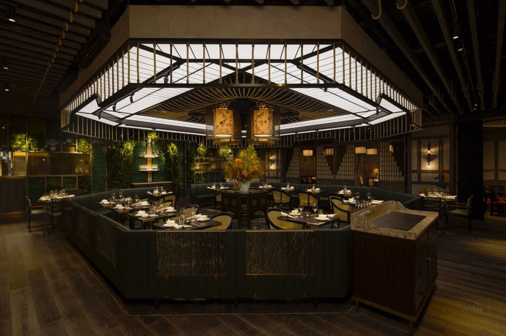 Elegant restaurant interior featuring ambient lighting and dark wood flooring. Central seating area with set tables, surrounded by green plants and decorative panels. Cozy and sophisticated atmosphere.