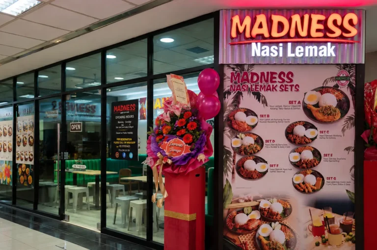 Madness Nasi Lemak restaurant storefront featuring a large menu board of nasi lemak sets and a congratulatory flower stand at the entrance