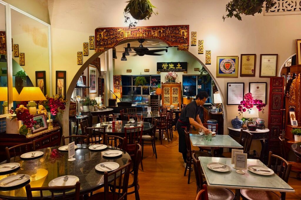 Cozy Asian restaurant interior with ornate decor, warm lighting, and wooden furniture. A staff member sets a table. Vibrant flowers enhance ambiance.