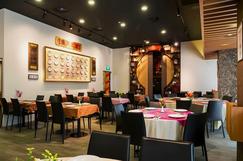 Modern restaurant interior with round tables, black chairs, and colorful tablecloths. Wall art features decorative plates. Warm, inviting ambiance.