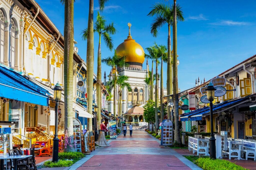 A vibrant street with colorful shops and tall palm trees leads to a grand mosque with a large golden dome under a clear blue sky, evoking a lively, cultural atmosphere.
