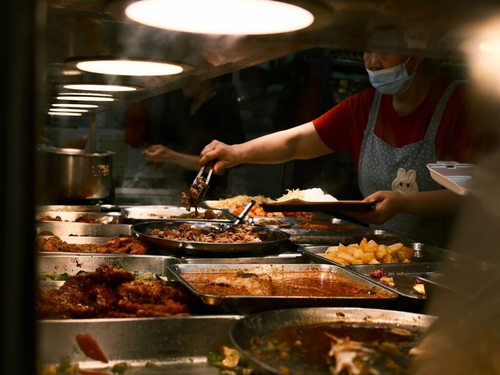 A food vendor wearing a mask and apron serves various dishes, including meats and vegetables, from steaming trays under bright overhead lights. The mood is bustling.