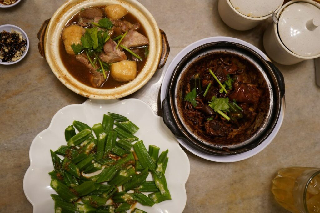 Top view of a dining table with diverse Asian dishes. A pot with meat and herbs, a claypot dish, and a plate of sautéed green vegetables. Cozy, inviting atmosphere.